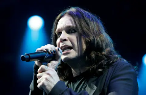 Getty Images Ozzy Osbourne has long, wavy dark hair and is holding a black microphone close to the mouth while performing on stage. The individual is wearing a black outfit, and the fingernails are painted black. A bright blue spotlight shines in the background, creating a dramatic concert atmosphere with dark surroundings and focused stage lighting.