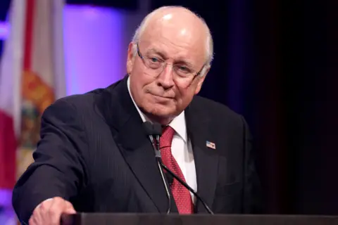 Getty Images Dick Cheney is standing at a podium, wearing a dark pinstripe suit with a white dress shirt and a red patterned tie. A small American flag pin is visible on the left lapel. He is speaking into a microphone, and part of a flag with red and white stripes is visible in the background, along with purple stage lighting. The setting appears to be a formal event or conference.