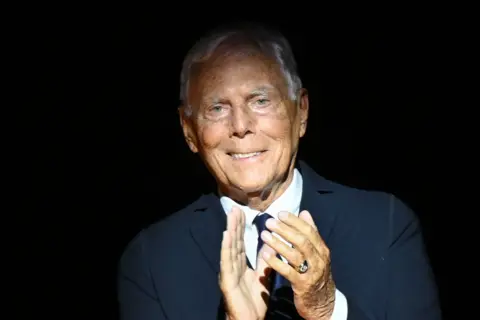 Getty Images Giorgio Armani is wearing a dark suit jacket over a white dress shirt, paired with a dark tie. His hands are positioned in front, mid-clap, with a ring visible on one finger. The background is completely black, creating a dramatic contrast and emphasizing the formal attire and gesture.