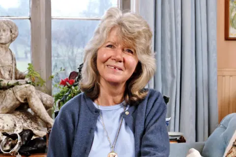 Getty Images Jilly Cooper is seated indoors on a light-coloured sofa, wearing a light blue top layered with a blue cardigan and a pendant necklace. Behind her, there is a window with light blue curtains, showing a view of trees outside. To the left, a decorative statue and a vase with red flowers are placed on a table, adding an artistic touch to the setting. 