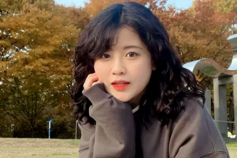 Instagram Baek Se-hee has shoulder-length curly dark hair and is sitting outdoors on grass, wearing a loose, long-sleeved gray sweatshirt. The background features autumn-coloured trees with shades of orange and yellow, and a metallic curved structure partially visible on the right side. She appears to be in a park or open recreational area during autumn.