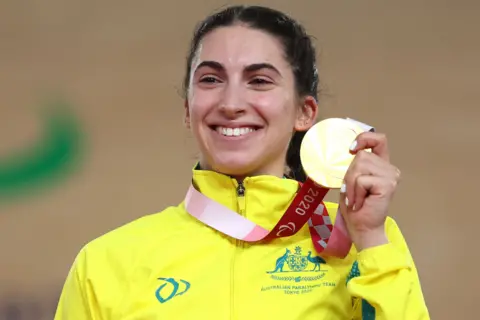 Getty Images Paige Greco wearing a bright yellow jacket with the Australian Paralympic Team logo and the text “Tokyo 2020” is holding up a gold medal attached to a red and white ribbon. The medal is prominently displayed in the foreground, and the background is softly blurred, suggesting an indoor sports venue or podium setting.