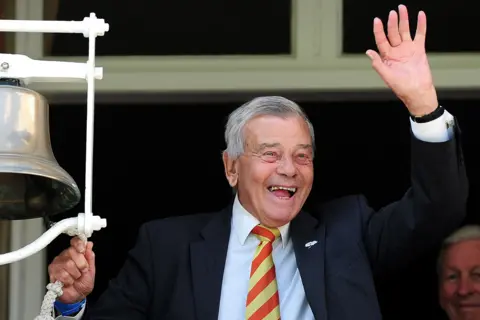 Getty Images Dickie Bird is wearing a dark suit jacket, a white dress shirt, and a striped tie in red and yellow is standing next to a large silver bell mounted on a white frame. One hand is holding the bell’s rope, while the other hand is raised in a waving gesture. The background shows part of a building structure with a window frame.
