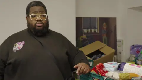 BBC Solomon Smith leans one arm on a green crate of donated food.