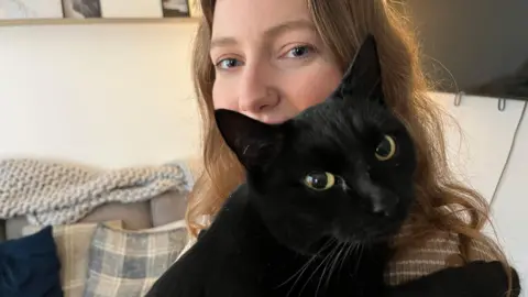 BBC A woman with long brown hair holding her black cat looking at the camera