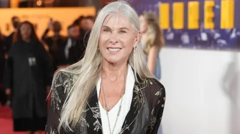 PA Media Sharron Davies wearing a sheer black jacket with metallic floral patterns over a white blouse, paired with light-colored trousers. The individual is standing on a red carpet at an event, holding a light handbag. 