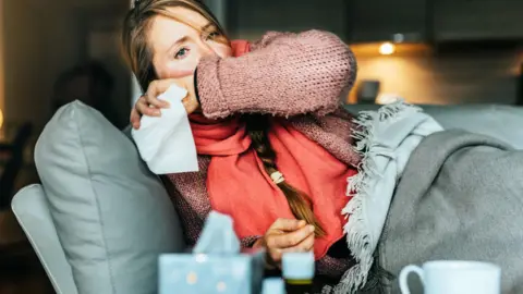Getty Images A woman with flushed cheeks lies on a couch covered in blankets. She covers her mouth, apparently coughing, and has a tissue in her hand. She has long blonde hair in a pleat and is wearing a pink jumper and a coral scarf. In the foreground of the shot is a box of tissues, a mug and two medicine bottles.