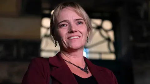 PA Media A portrait of a woman, shown from the shoulders up, with a dark background and soft lighting. She has medium-length blonde hair that is neatly styled.