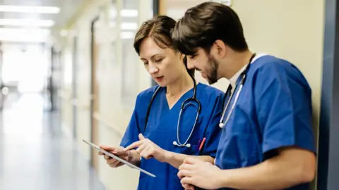 Getty Images A male and female doctor, both wearing blue scrubs, standing in an empty hospital hallway examining an iPad