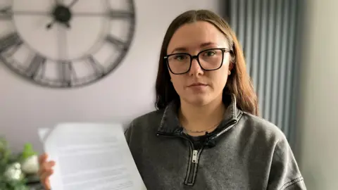 Oli Constable/BBC A woman with brown, straight hair looks at the camera while holding a piece of paper in her right hand