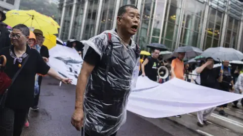 Getty Images Jimmy Lai holds a banner and is wrapped in plastic overlay as he marches in the rain along Queen's Road Central during a protest in the Central district of Hong Kong, China, on Sunday, Aug. 18, 2019.