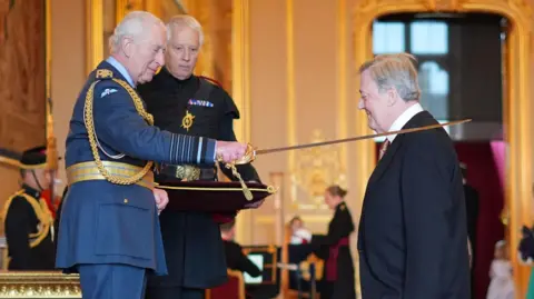 PA Media The actor Stephen Fry kneels as King Charles places a sword on his left shoulder during his investiture at Windsor Castle on 25 March 2025.