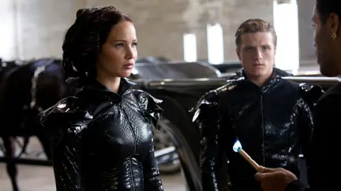 Lionsgate Still from Hunger Games: Mockingjay Part 2 shows Jennifer Lawrence and Josh Hutcherson in costume, being addressed by a third character just off-screen.