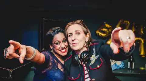 Pretty Ugly Two women - Aarti Joshi and Kirsty Wark - standing behind the decks in a club. Both are pointing towards the camera, with headphones round the neck of Kirsty Wark.