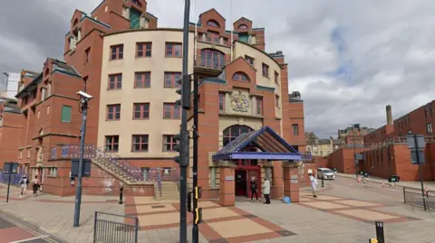 Google A large, stately building dominates the scene, constructed with alternating bands of red brick and beige stone. Its façade features tall arched windows framed by decorative molding, and the roofline is punctuated by steep gables and dormers. Above the central entrance, a crest or emblem is prominently displayed.