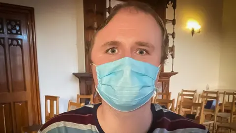 David White A head shot of David White looking right into the camera. He is wearing a blue hospital mask and a round-neck stripy shirt. His eye are blue and he has receding light brown hair. The right side of his head has a large indent on the top. 