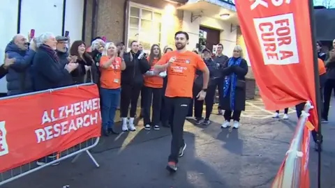 BBC A man in an orange t-shirt has his arms raised in a walking stance as he sets off on a charity challenge. He has black hair and a dark, short beard. A crowd is clapping and cheering behind him, and orange banners display logos for Alzheimer's Research UK.