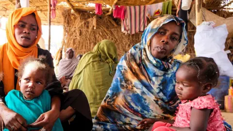 Reuters Two women wearing headscarves sit side by side, holding young children in their arms.