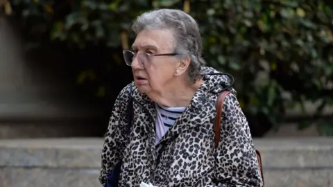 Spindrift Patricia Robertson, a 77-year-old woman with short grey hair and clear-rimmed glasses, is walking right to left. She is wearing a leopard skin hooded jacket and a  light and dark blue striped shirt underneath. She has a brown leather bag slung over her shoulder.