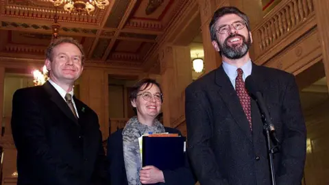 Getty Images Gerry Adams, Martin McGuinness and Bairbre de Brun standing together smiling. Gerry Adams has a microphone in front of him. He is wearing glasses, a suit, light blue shirt and red patterned tie. Bairbre de Brun is holding a file and is wearing a grey patterned scarf, navy jacket and glasses. Martin McGuinness has a black suit on with a white shirt and gold patterned tie.