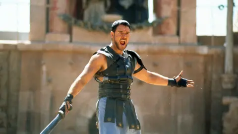 Getty Images Russell Crowe is dressed like a Gladiator in a leather chest guard and he is wielding a sword. He is shouting and is arms are raised. 