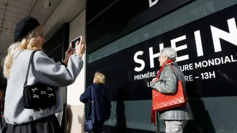 Getty Images A woman takes a picture of a Shein banner at a Paris department store as an elderly woman carrying a red handbag walks by.
