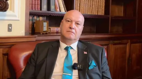 BBC Ian Cooper sits in a maroon leather chair, with wooden panelling and a recessed bookcase behind him. He is wearing a white shirt, dark grey suit jacket, with a union flag pin badge and a clip microphone on the lapel, and a teal blue tie. He is looking into the camera with a serious expression.