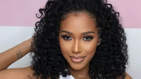 Aiesha Barrett A woman with black curly hair smiles at the camera in a relaxed pose, with her head resting one on hand. She has a glamorous look, with well-manicured eyebrows, a glossy lip and long eyelashes.