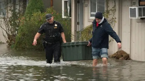 Reuters Deputy Sheriff Kalani Apilado helps Brandon Phasith carry belongings while evacuating amidst rising floodwater, as an atmospheric river brings rain and flooding to the Pacific Northwest, in Sultan, Washington
