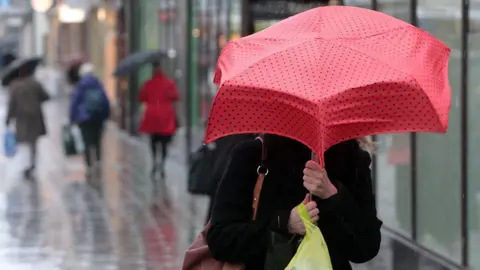 Getty Images A woman holding a red umbrella with black polka dots on it walks into the wind. Her face is covered by the umbrella and she is wearing a black coat with a brown handbag on her shoulder and a yellow carrier bag in her hand. She is on a shopping street, which is wet with rain, and blurred people in the background are also seen holding umbrellas. 