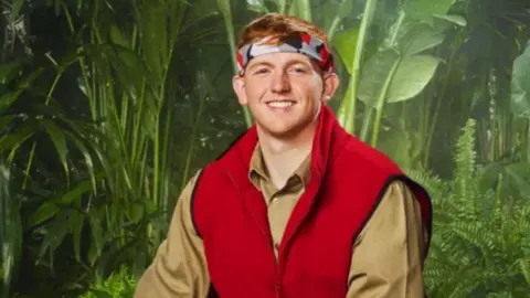 ITV Angry Ginge in a promotional picture for I'm a Celebrity. He wears a red gilet, camo bandana and is smiling.