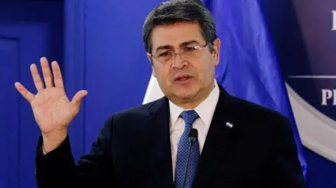 Reuters Honduras' President Juan Orlando Hernandez speaks at the Presidential House in Tegucigalpa, Honduras in January 2020. He is wearing a suit and navy blue tie, behind him is a royal blue backdrop. He is gesturing with his right hand. 