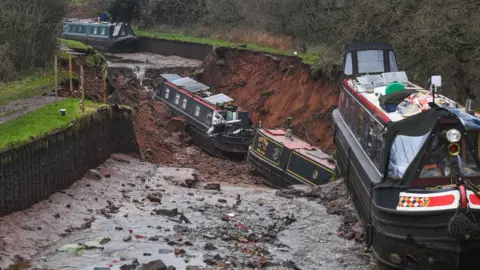 PA Media Two canal boats in a large hole of a drained canal. On earth above them is another boat hanging over the edge of the hole