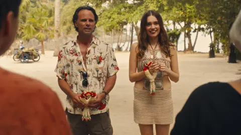 Warner Bros Discovery Walton Goggins and Aimee Lou Wood,  in costume, stand next to each other on a beach in a scene from the White Lotus