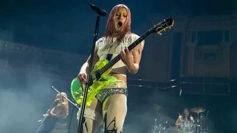 Getty Images A rock band performing onstage. Among them is their singer, wearing a white T-shirt and glittery silver hot pants, playing a neon green guitar. The band's drummer and another guitarist can be seen in the background