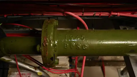 House of Commons A green pipe with the year 1945 embossed on the side. There are red wires behind the green pipe.