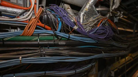House of Commons Several groups of different coloured wires running along a corridor. Some of the wires wrap around others or around one of the pipes