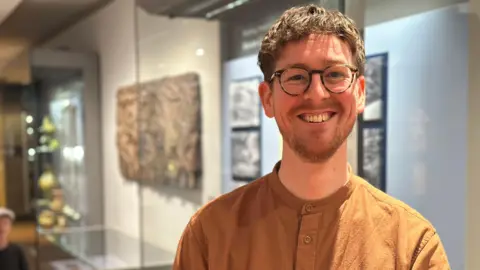 Luke Holmes has brown hair and glasses. He smiles at the camera while wearing a brown shirt. He's standing in a museum in front of glass cabinets.