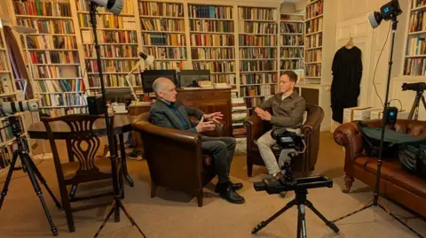 Prof Peter Wilson is sat in an office full of books opposite a journalist. They sit in leather arm chairs and around them are lights and cameras to record the interview.