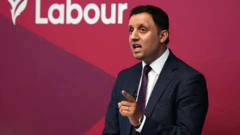 PA Media Anas Sarwar, who has short black hair, speaks with his left hand in front of him, with the index finger pointing. He is wearing a dark suit, purple tie and white shirt, and standing in front of a red Labour-branded wall 