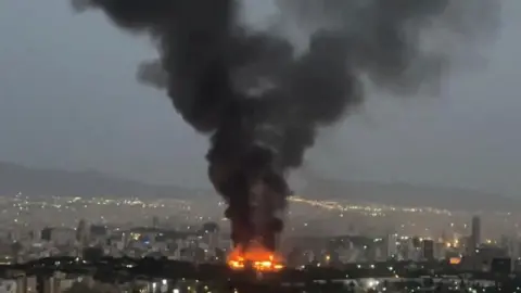 Reuters A fire burns in Tehran as a pillar of black smoke rises above the skyline (17/06/25)
