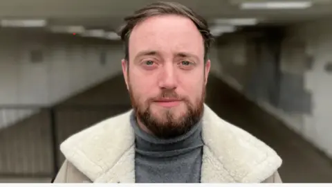 Jacob has a beard and is wearing a jumper and warm jacket