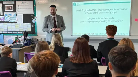 Teacher James Stainton, a man with short dark hair who is wearing a grey suit and burgundy tie, stands in a secondary school classroom giving a lesson on the risks of vaping. Nine young people are seen from behind, seated at desks and wearing school uniform. Text displayed on a white board states that according to NHS England, of 2024, "1 in 10 teenagers in secondary schools in the UK currently vape." The texts asks students to consider "Why is vaping so appealing to teenagers?" 
