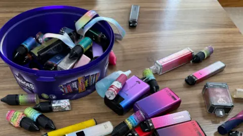 Several vapes lay scatted on the head teachers desk. Some are stored in a Heroes chocolates box