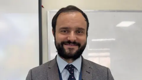 A man with dark hair and a dark beard and hazel eyes. He is wearing a grey suit jacket, light blue shift, navy tie with pink shapes on it. He is standing in front of a whiteboard with nothing written on it.