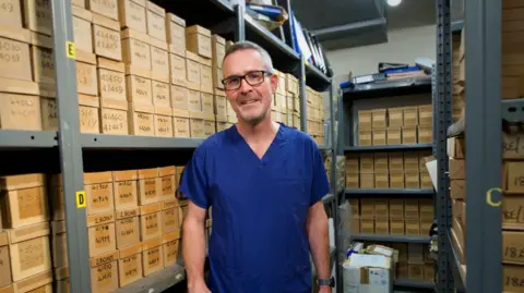 Man in blue scrubs and glasses stands in front of floor to ceiling shelves stacked with brown cardboard boxes.