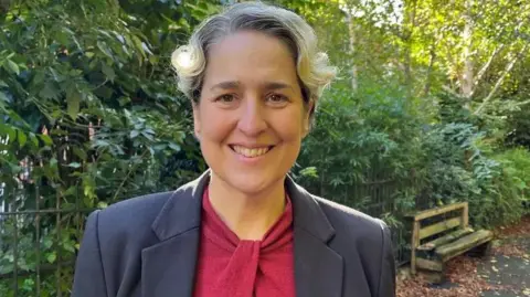 Caroline Woodley is pictured shoulders up smiling directly at the camera. She is wearing a read blouse and dark blazer. 
