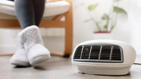 Getty Images Close up shot of a woman wearing thick grey tights and white slipper boots next to an electric heater. 