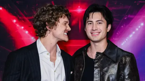 Getty Images Connor Storrie and Hudson Williams posing together in front of a red, purple and black backdrop, with Storrie pulling a comical grin at Williams