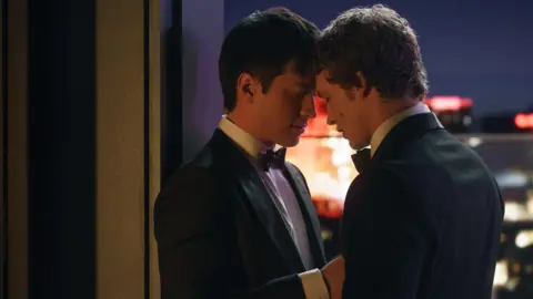 Sabrina Lantos/HBO Max Hudson Williams and Connor Storrie in a scene from Heated Rivalry, both wearing dinner jackets and bow ties, facing each other with their heads affectionately touching on a balcony at night